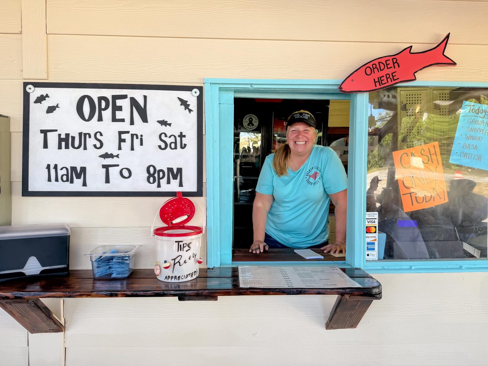 Owner Julie Cockerham smiling at the turquoise order window beneath the Open Thurs Fri Sat sign